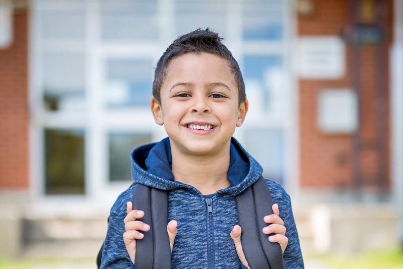 Elementary student smiling at camera and gripping shoulder straps of backpack while standing in front of school building