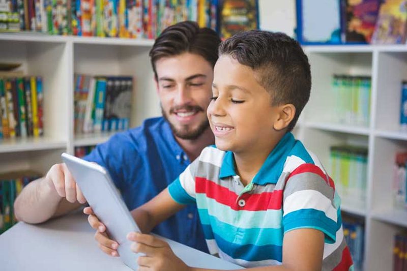 Elementary student smiling and holding tablet while teacher points at screen